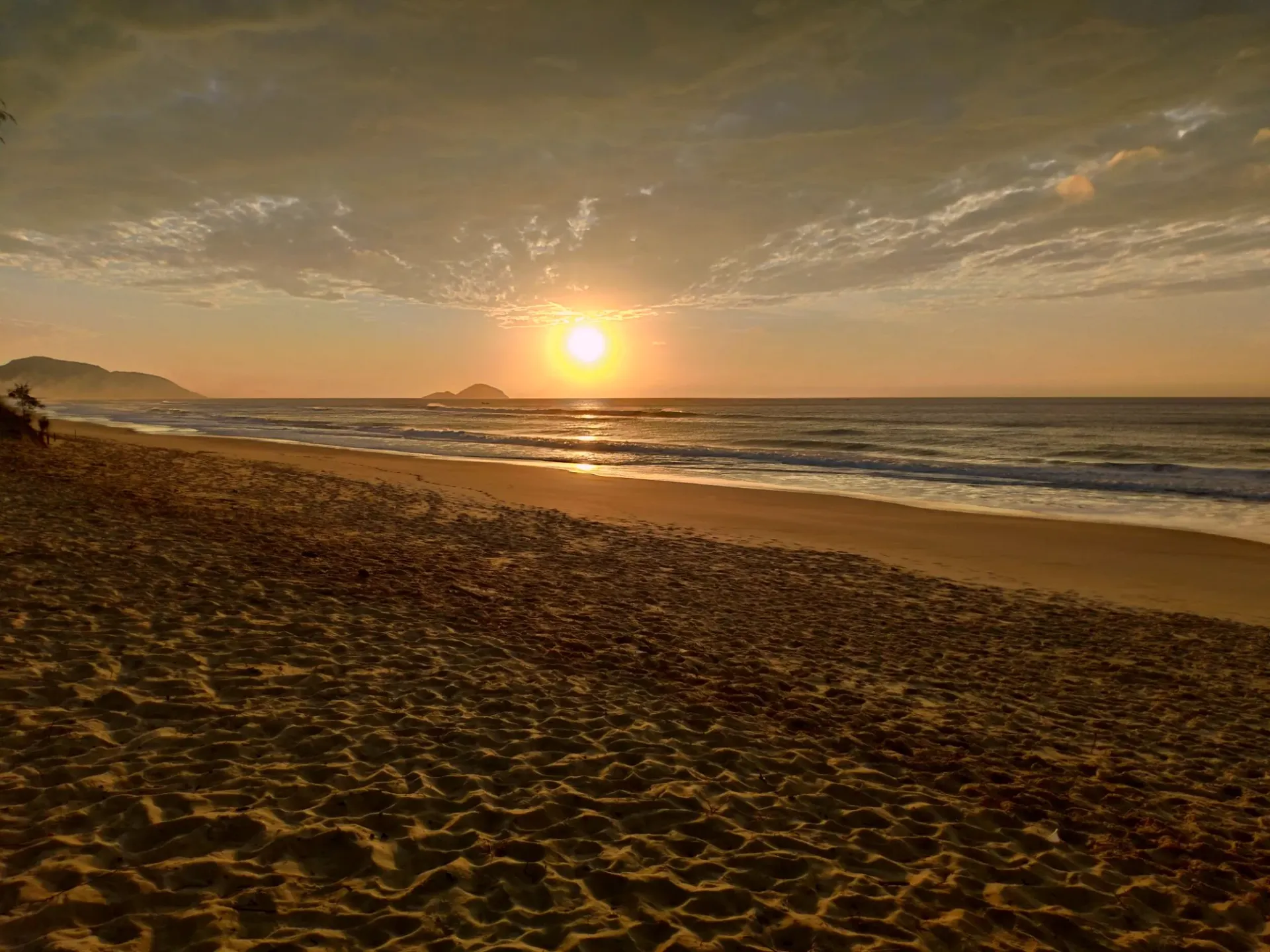 Sol nascendo sobre o mar na Praia Mole, com a areia em primeiro plano, um morro à esquerda e uma ilha no horizonte